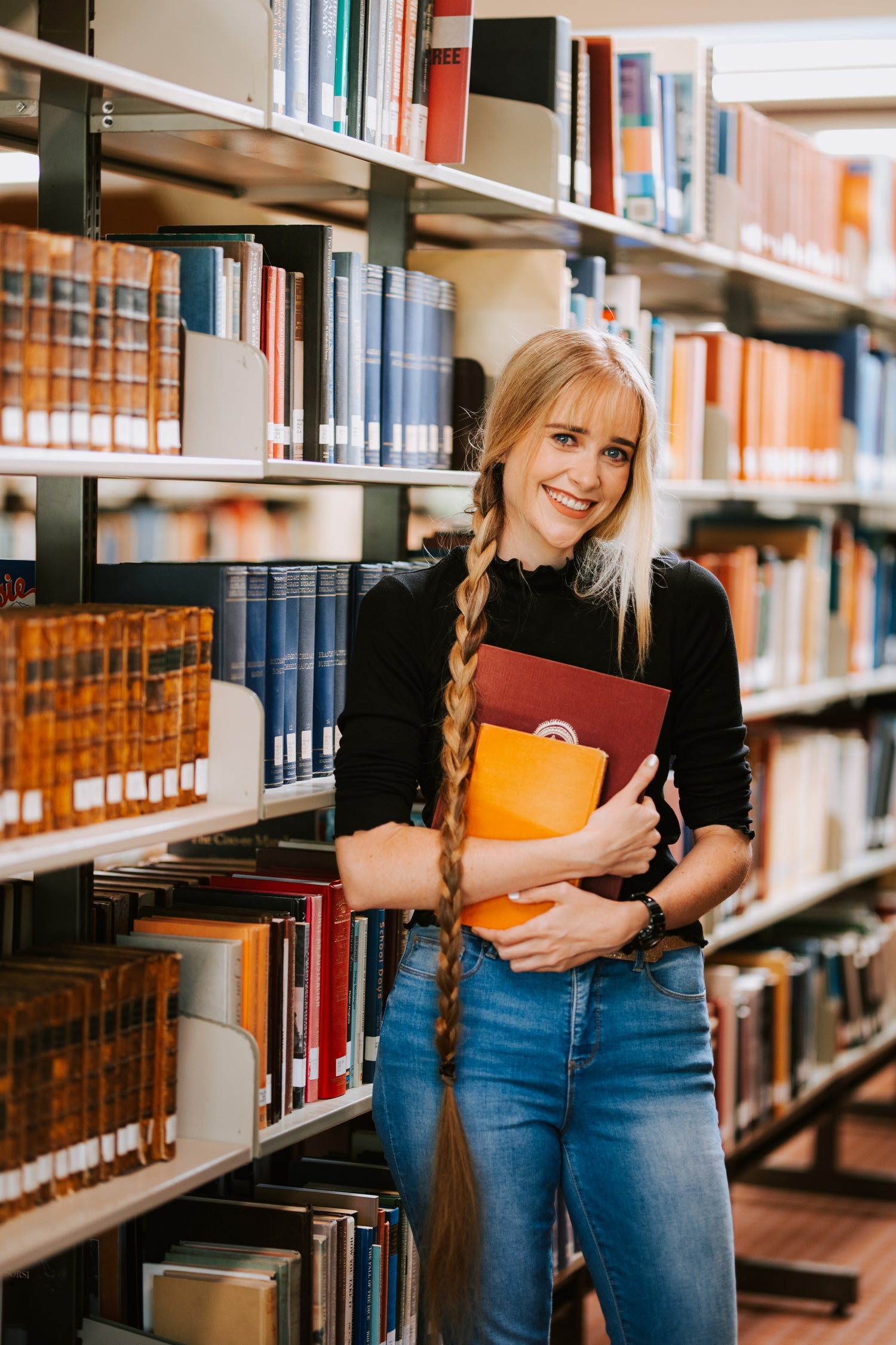 Toby Hendy standing in a library holding books. Her hair is in a braid that reaches to her knees. She is smiling.