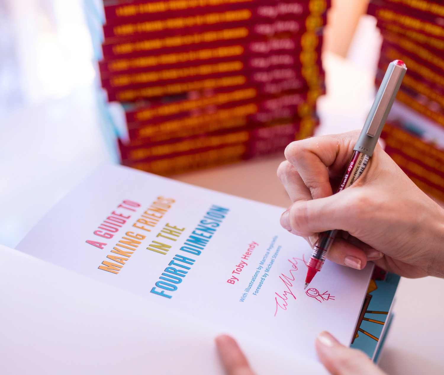 Person signing a book titled 'A Guide to Making Friends in the Fourth Dimension' with a pen.