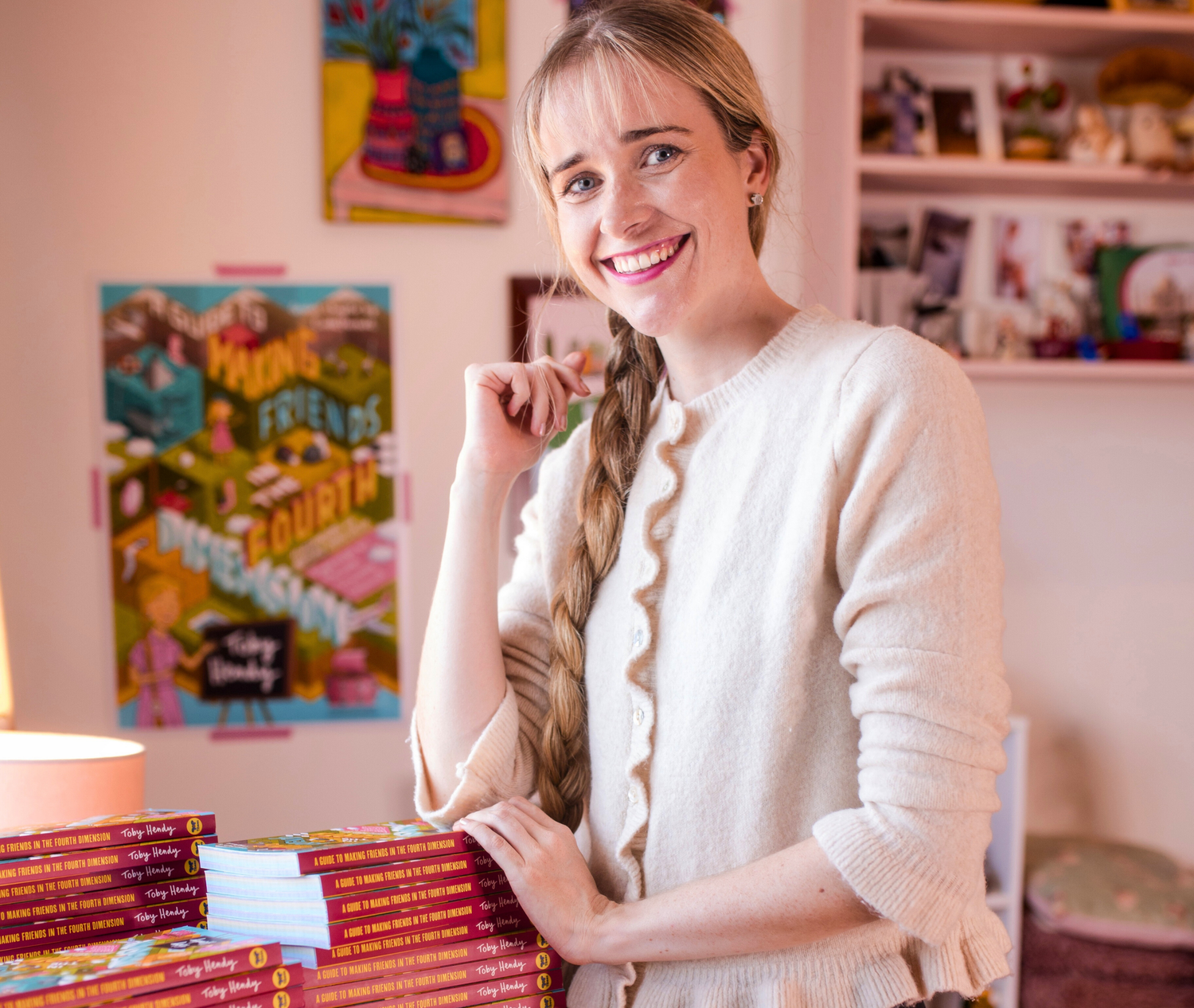 Woman in a white sweater standing next to a stack of books in a room with colorful posters on the wall.